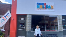 A woman wearing a blue skirt and white cardigan stands in front of a building painted in bright orange on the side and the words "Feed Halifax" on the face of the building. 