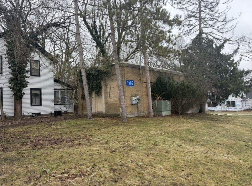 On a patchy, muddy lawn, a small brick building sits surrounded by trees and covered in moss. The building has no windows, just a small door and a blue plaque with the numbers 1509
