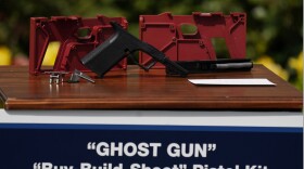 A red polymer frame is displayed on a table with pieces of a pistol in front of it. A sign below the display reads "GHOST GUN 'Buy-Build-Shoot' Pistol Kit."