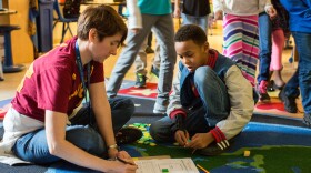 A teacher reviews a student’s completed work in a third-grade classroom as students rotate to new math stations.