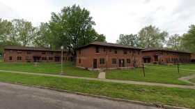 Three two-story red brick apartment buildings with brown roofs sit on a grassy lawn, surrounded by trees and sidewalks, under a cloudy sky.