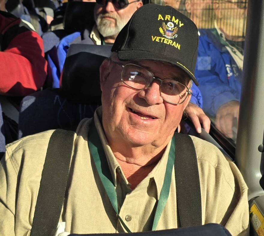 An elderly man is seated on a bus, smiling at the camera. He's wearing glasses and a hat that says "Army Veteran."