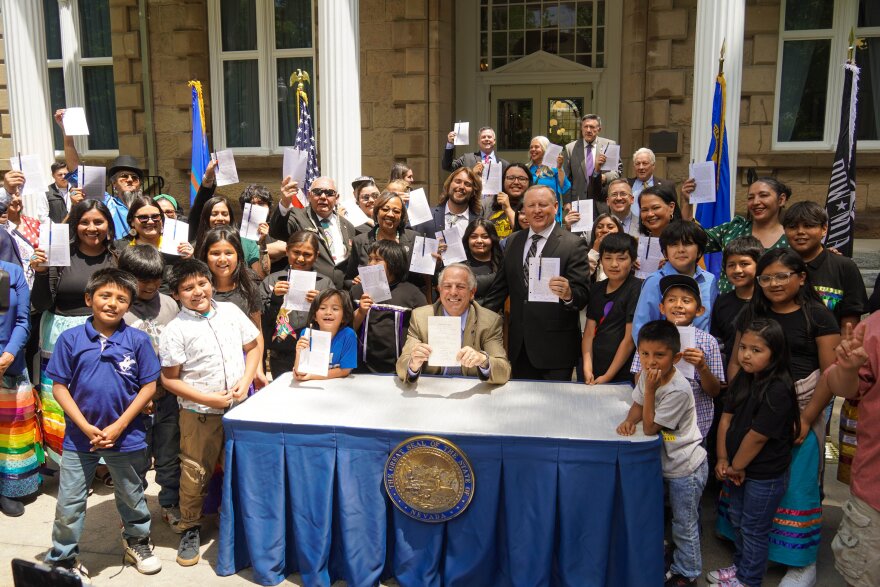 A group of about 30 people are standing around a man sitting at a table and holding up a piece of paper.