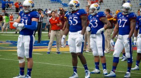 Medium range photo of four University of Kansas football players standing on the field wearing their home uniforms of white helmets with the Jayhawk mascot on the side, dark blue shirts with the word Kansas and their number on the front, white pants with wide dark blue stripe down the side, blue socks and white cleats (shoes) with dark blue laces.