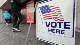A large "Vote Here" sign with an American flag is in foreground. Two people can be seen in background walking into the entrance to the National World War I Museum and Memorial entrance.