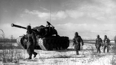 Four US soliders march through the snow next to a tank. 