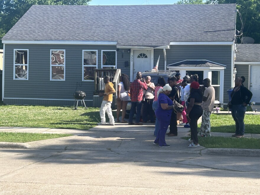 The image is of a gray house during the day. A group of people stand in the driveway of the house. 