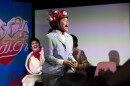 A person wearing a gray suit and red hat, dressed as Pee Wee Herman, peforms on stage holding a rubics cube. Behind them is a person dressed in a white jumpsuit as Elvis. 