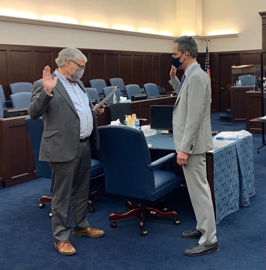 two men wearing gray suits and masks being sworn in to a role inside a courtroom bedecked with brown wood and blue carpets and chairs