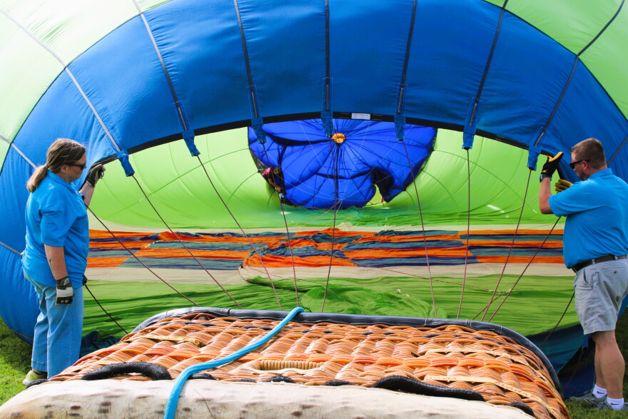 Two people in blue shirts hold open the bottom of a deflated hot air balloon, preparing to inflate it. The balloon’s colorful fabric is spread out on the ground, with the basket in the foreground.
