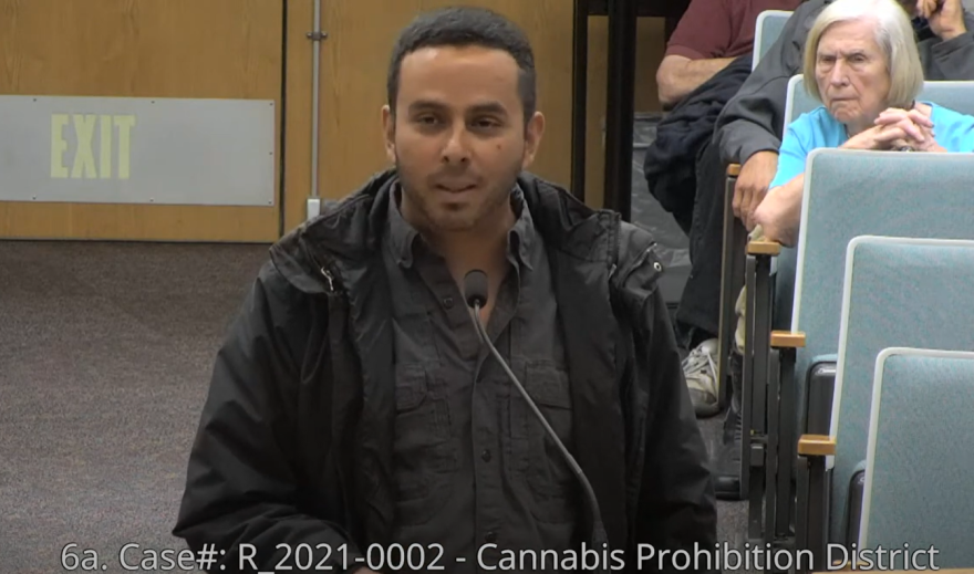 A man in dark clothing speaks into a microphone as a woman scowls in the background.