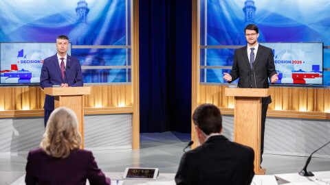 Two men in suits stand at podiums facing a man and woman sitting at a table in a TV studio
