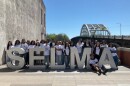A school group stands behind a silver sign reading, 'Selma.' The Edmund Pettus Bridge is behind them.