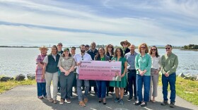 16 people stand together, smiling at the camera and holding a large check that reads "One Million Ninety Two Thousand Dollars." They're standing in front of a shoreline, with blue skies covered in large streaks of clouds.