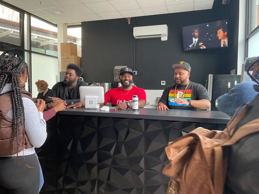 Three men stand behind a black counter with a POS system. From left to right is James Dixon in a black shirt, David Dixon in a red shirt and black hat, and Mario Savage in a black shirt with a black hat. Customers are in the forefront of the frame.