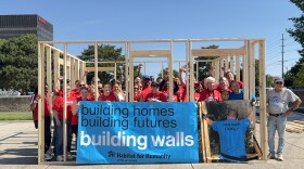 Volunteers in red T-shirts stand inside the frame of a square house, holding a banner that reads "building homes, building futures, building walls."