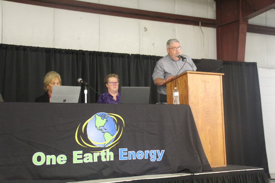 A business executive speaks at a podium next to a table set up for a public meeting. 