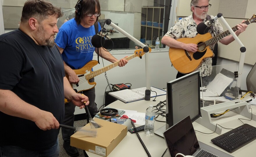  Three people stand in a radio studio, the one on the far left plays a box with drum brushes, the one in the middle sings while playing bass guitar, and the person on the far right plays an acoustic guitar