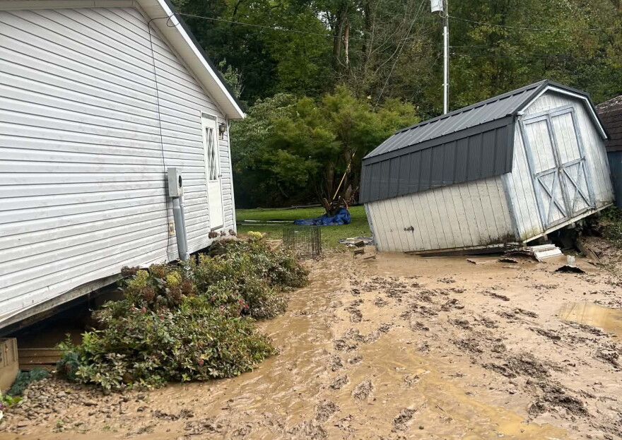 A storage shed that has been knocked over by flood waters sits in a muddy yard.