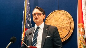 A man in a dark suit and tie stands in front of a blue wall with an American and State flag behind him. 