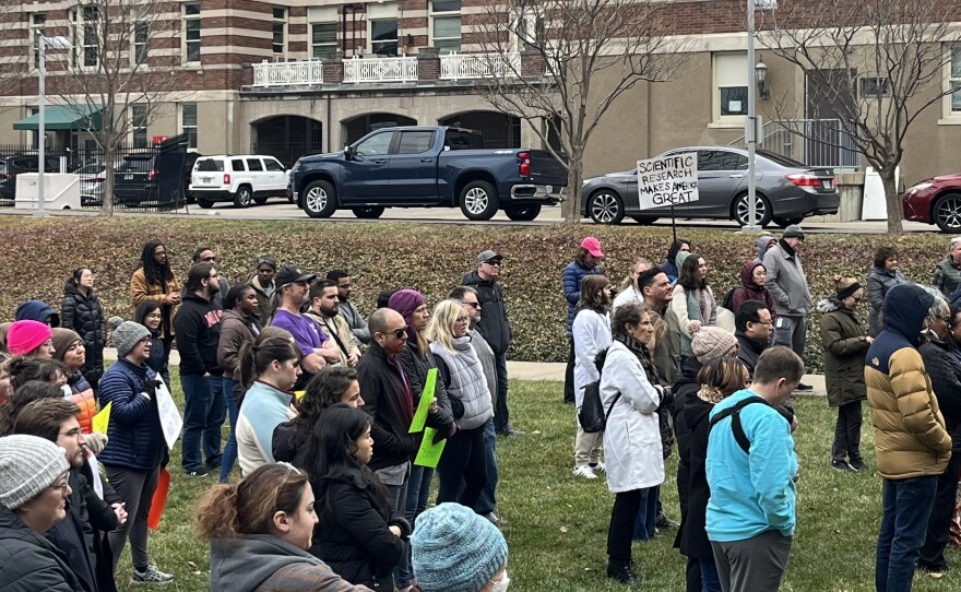 People wearing winter coats and hats and holding protest signs stand in a park.