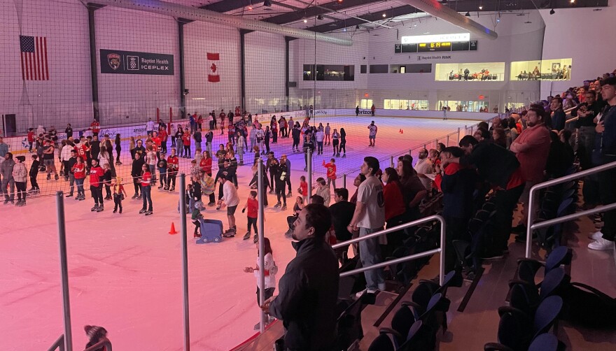 Fans on an ice rink and in the stands look to the left of frame as a dull red light bathes their faces