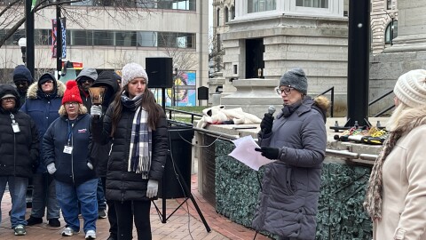 A crowd of singers stand behind a woman holding a bell who is next to two other women, reading from a list of names that recognize lives lost in 2025 after experiencing homelessness.