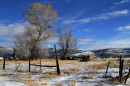 An old, worn-down shed sits in a field with yellow grass and snow. There are old wood fences in the foreground and a mountain range in the background.