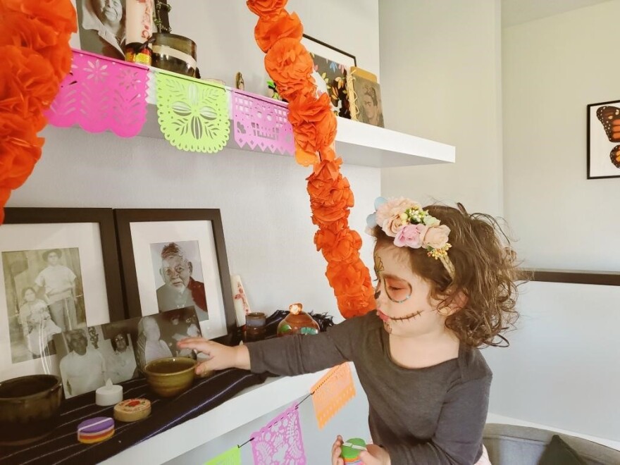 Verónica Guevara pintó la cara de su hija Emma como una calavera de azúcar mientras trabajaba en su altar para el Día de los Muertos. Guevara dijo que Emma, de 2 años, intentó quitar los dulces del altar. “Yo le dije, ‘no, eso es de mi abuela. Esos son de tu bisabuelo”, dijo Guevara. “Ella los dejó y luego me pidió que le diera sus propios dulces.”