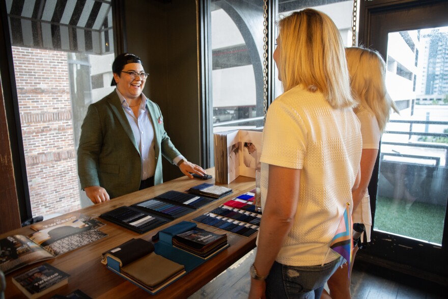 A person wearing a backward baseball cap and a green suit stands behind a table with clothing swatches. A blonde haired couple looks through the samples. 