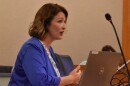 A woman with wavy brown hair wearing a blue blazer speaks in a council chamber