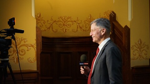 Wyoming Governor Mark Gordon stands before a TV news reporter at the state capitol in Cheyenne, Wyoming. Gordon is looking toward frame left, and is framed by yellow walls in the background.