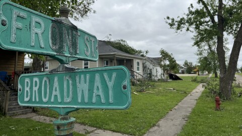 A street sign showing the intersection of Front and Broadway in Slater, MO, with downed tree limbs damaging houses in the background.
