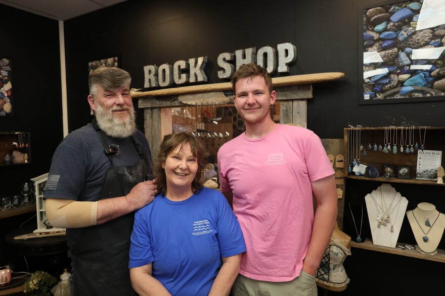 An adult man, an adult women, and a male teenager smile for the camera. Behind them is a metal sign that says “Rock Shop” and displays of rock-based jewelery and photos. 