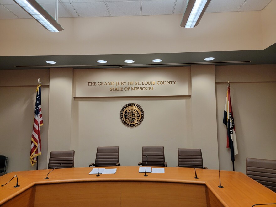 Brown high-backed chairs sit around a tan panel desk with microphones. The American flag sits to the left of the seal of the state of Missouri and the Missouri flag to the right. The words the grand jury of St., Louis County, state of Missouri, are written on the wall above the seal.