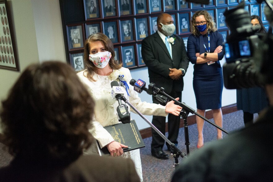 Assemblywoman Teresa Benitez-Thompson stands in front of a group of microphones answering press questions. She is wearing a white floral dress and mask and is holding a notebook in front of her.