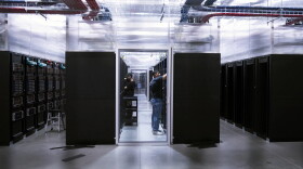 Two people work inside a glass-walled server room surrounded by tall black server racks in a bright, modern data center with overhead cabling—an environment where sustainability is prioritized through the use of hydropower to efficiently run the facility.