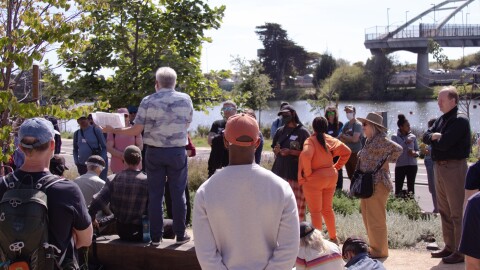 A group of about 30 people standing outside in the sun in front of the water at Berkeley Aquatic Park. Most people's backs are to the camera, but a few are facing the camera, some people are sitting in the dirt. Their heads are cocked and you can tell that they are listening to Wholly H2O tour guide Chris James, who is standing on a bench so people can hear him more clearly. James has a full head of white hair and is wearing a white and blue button down shirt, and blue jeans. He is holding several pieces of paper in his left hand. Many people are wearing hats, and sunglasses. In the background, you can see a bridge, small oak trees, willow, and sage bushes. 