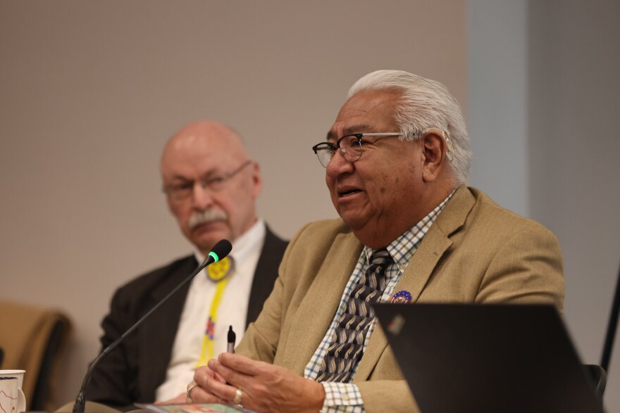 A man with white hair who is wearing a patterned tie and tan suit jacket speaks into a microphone.