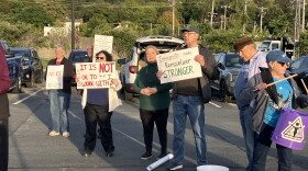 Several protesters are standing in a row in a parking lot. Most of them are holding posters, some of which say "It is NOT ok to with ICE" and "Immigrants make Rensselaer STRONGER." One woman is holding drum sticks and has a plastic bucket hanging from a strap around her neck. 