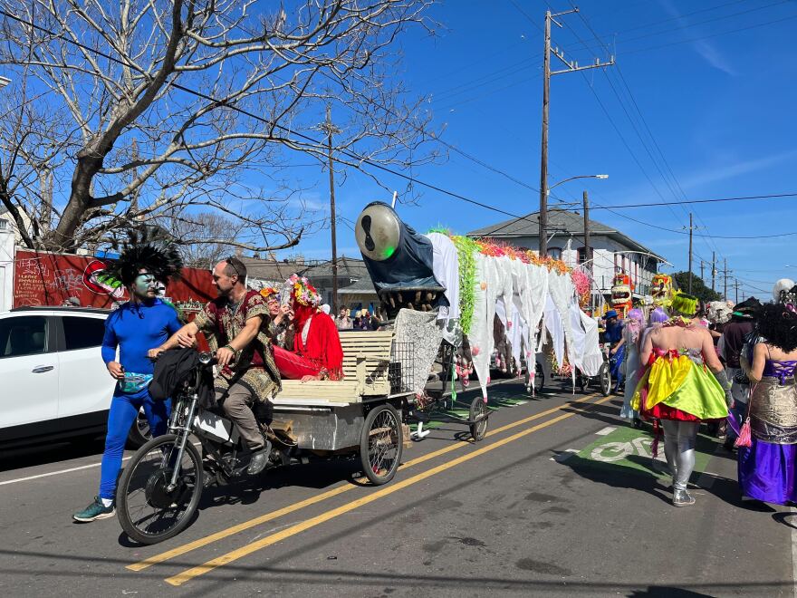 A man on a bicycle tows a long silver float draped in white cloth down the street during Mardi Gras.