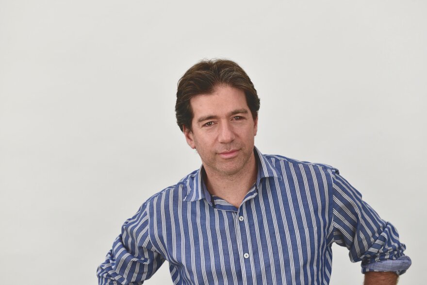 Composer David Shapiro, in a blue shirt with white stripes, looks towards the camera with a neutral expression, and with arms on his hips just out of view.