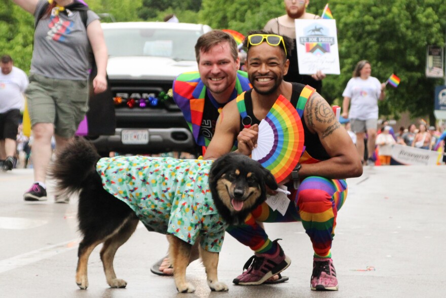 Two men in rainbow-striped outfits pose with their dog, who is wearing a tropical shirt.