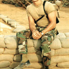 Miles Burdine in 1983 in military fatigues sits on sandbag wall