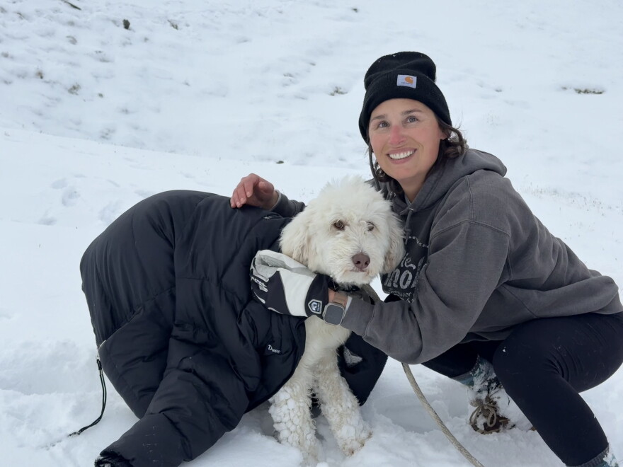 A woman kneels next to an off-white dog wrapped in a black puffy coat on a snowy background.