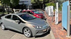A row of electric cars, plugged into charging stations. Office buildings in the background.