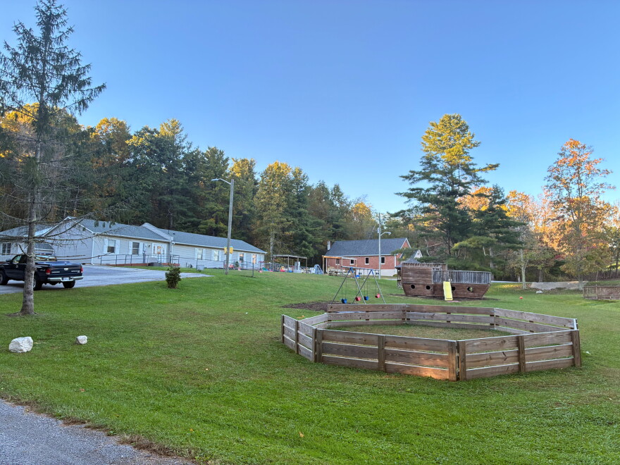 A green mowed yard with a swing-set, a wooden ship with a slide, and a fenced in area, with several building in the background. 