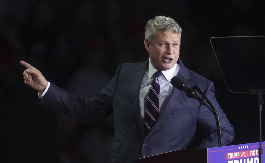 Man in dark suit and dark tie with lighter stripes gestures and smiles while pointing a finger and standing at a lectern with a sign that says "Trump will fix it, Trump Vance" (it cuts off at the edge of the frame)