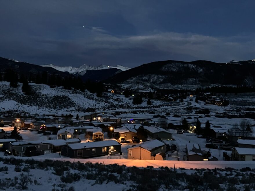 A neighborhood at night with some lights on and snowy hills in the distance.