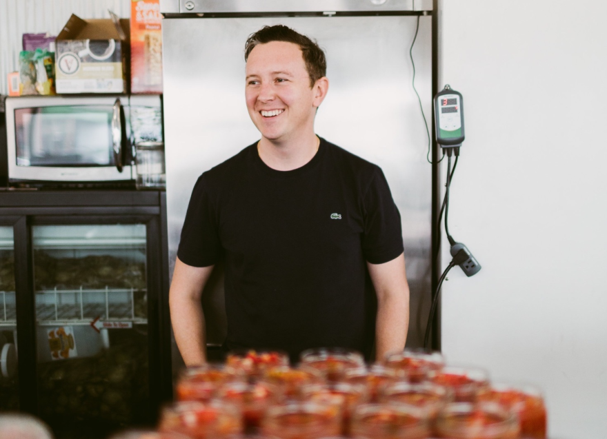 A man in a black t-shirt stands in front of several jars. Kansas City Canning Company owner Tim Tuohy. He learned about canning as a child from neighbors. Tuohy transitioned into culinary arts after losing his job as a teacher in 2009 during the recession.